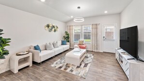 A modern living room with a sofa, coffee table, TV, and a large window. - Brand-New Home - Near Fort Sam & Randolph - Safe (Converse)