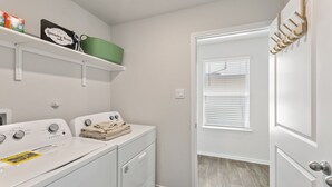 A laundry room with two washers and dryers, a shelf with laundry supplies, and a door with a coat rack. - Brand-New Home - Near Fort Sam & Randolph - Safe (Converse)