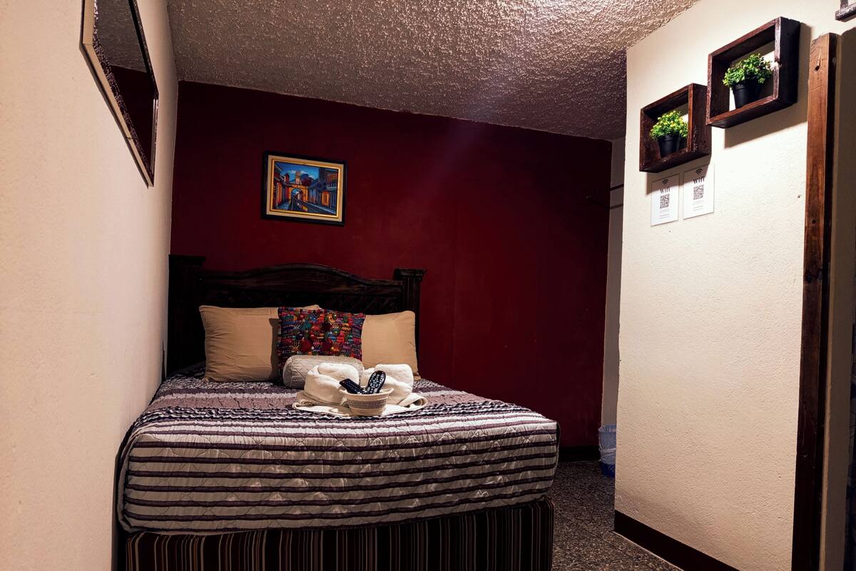 Un dormitorio con cama, almohadas, una colcha, un control remoto y un cuadro en la pared.