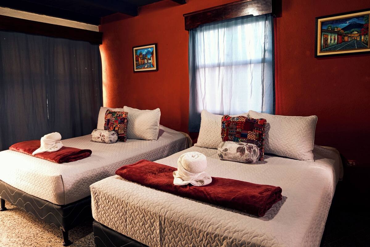 Habitación de hotel con dos camas, cada una con una manta roja y una almohada. Hay una ventana con cortina, un cuadro en la pared y suelo alfombrado.