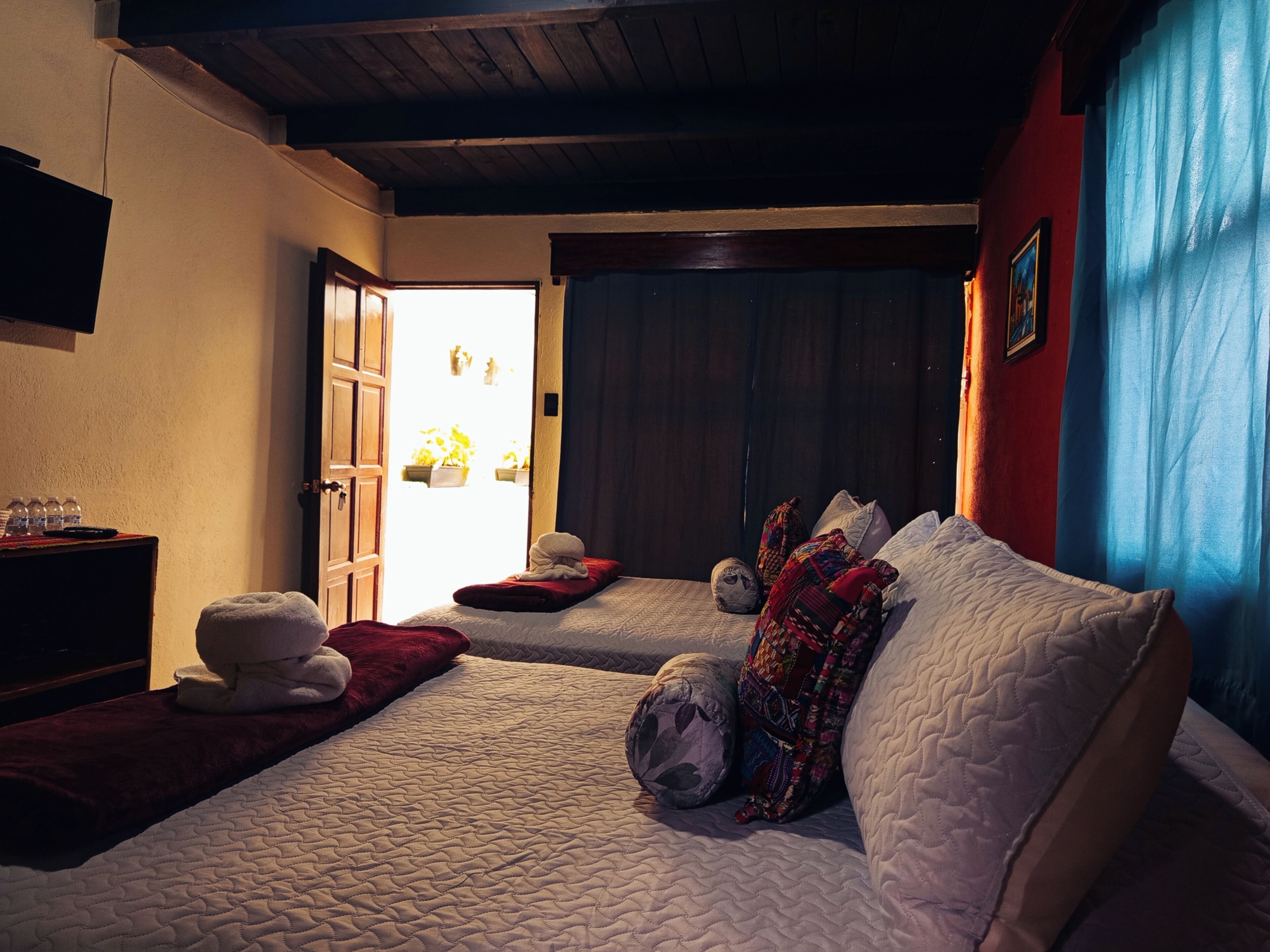 A hotel room with a bed, a small table with a towel, a wooden door, and a view of the outside.