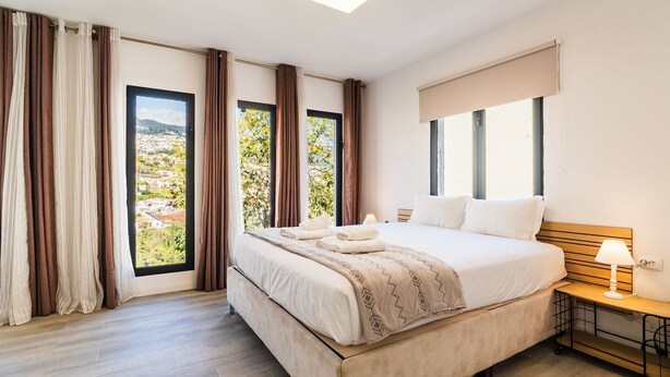 Un dormitorio con cama, mesita de noche, lámpara y vistas a un paisaje urbano a través de amplias ventanas.