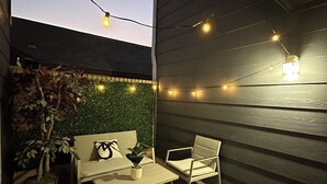 Eine Terrasse im Freien mit Holzdeck, einem weißen Sofa, einem weißen Sessel, einem kleinen Tisch und Lichterketten.