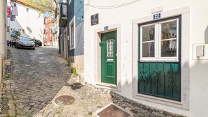 A narrow cobblestone street with a green door and window, a parked car, and a building with a sign indicating 'Rua das Escolas Gerais.' - GuestReady - Charming duplex in the Alfama heart (Lisbon)