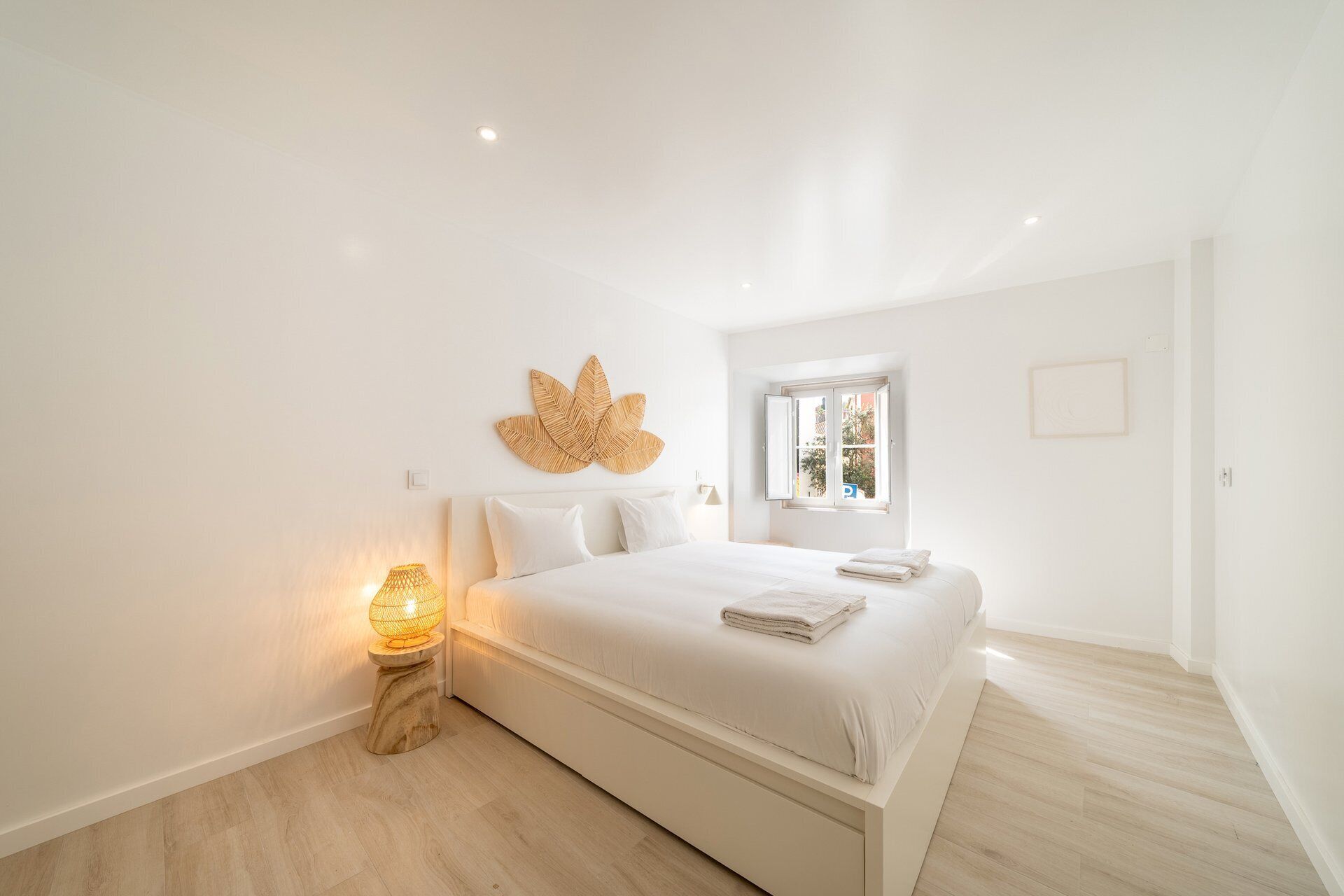 A minimalist bedroom with a bed, a wall-mounted leaf decoration, a bedside lamp, and a window with a view.