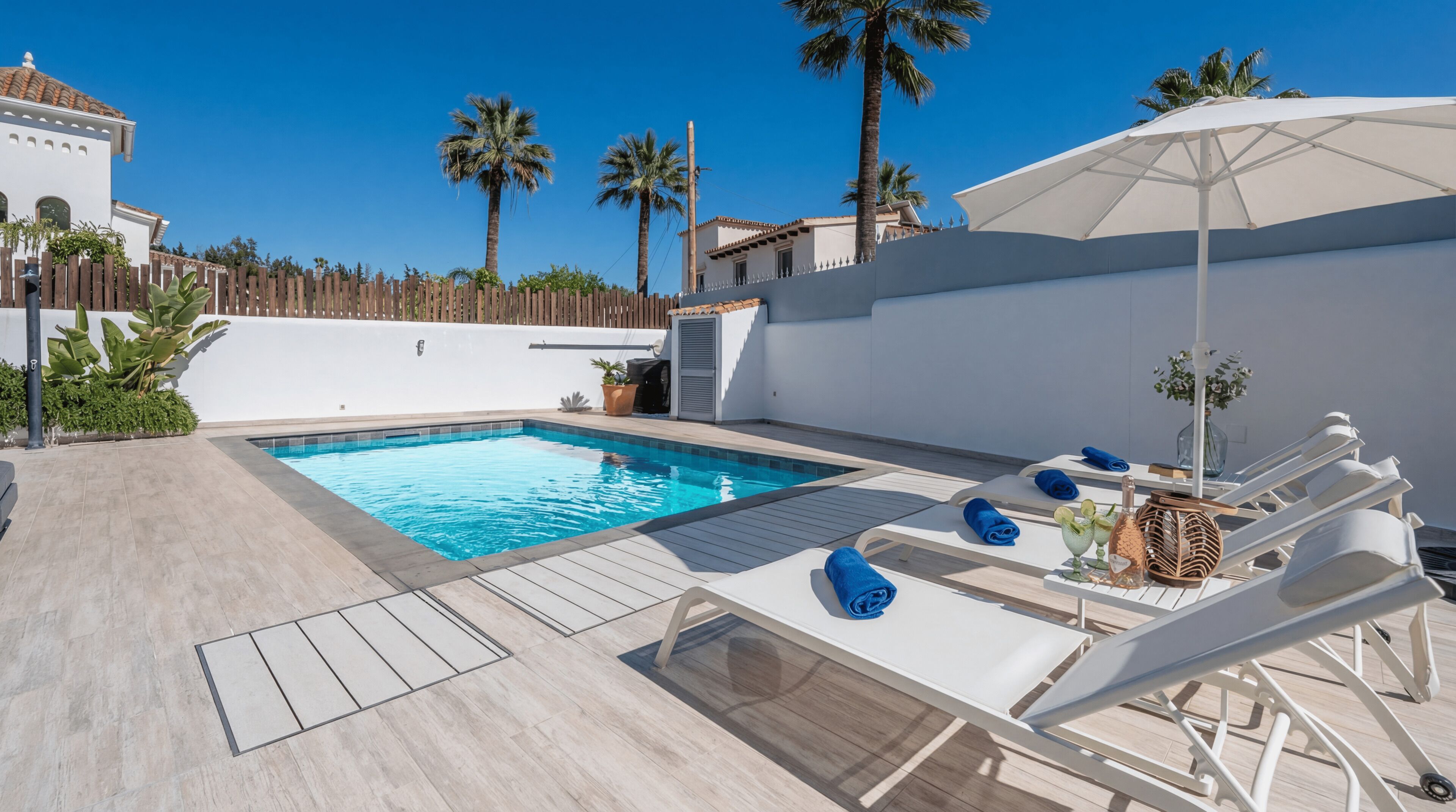 A swimming pool with lounge chairs and an umbrella, surrounded by palm trees and a white building.