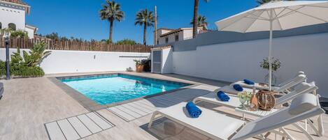 A swimming pool with lounge chairs and an umbrella, surrounded by palm trees and a white building.