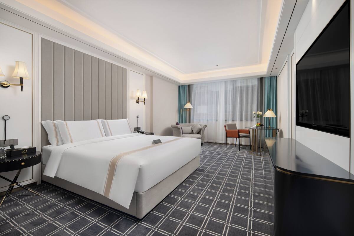 A hotel room with a large bed, a desk with a chair, a TV, and a small table with a lamp.