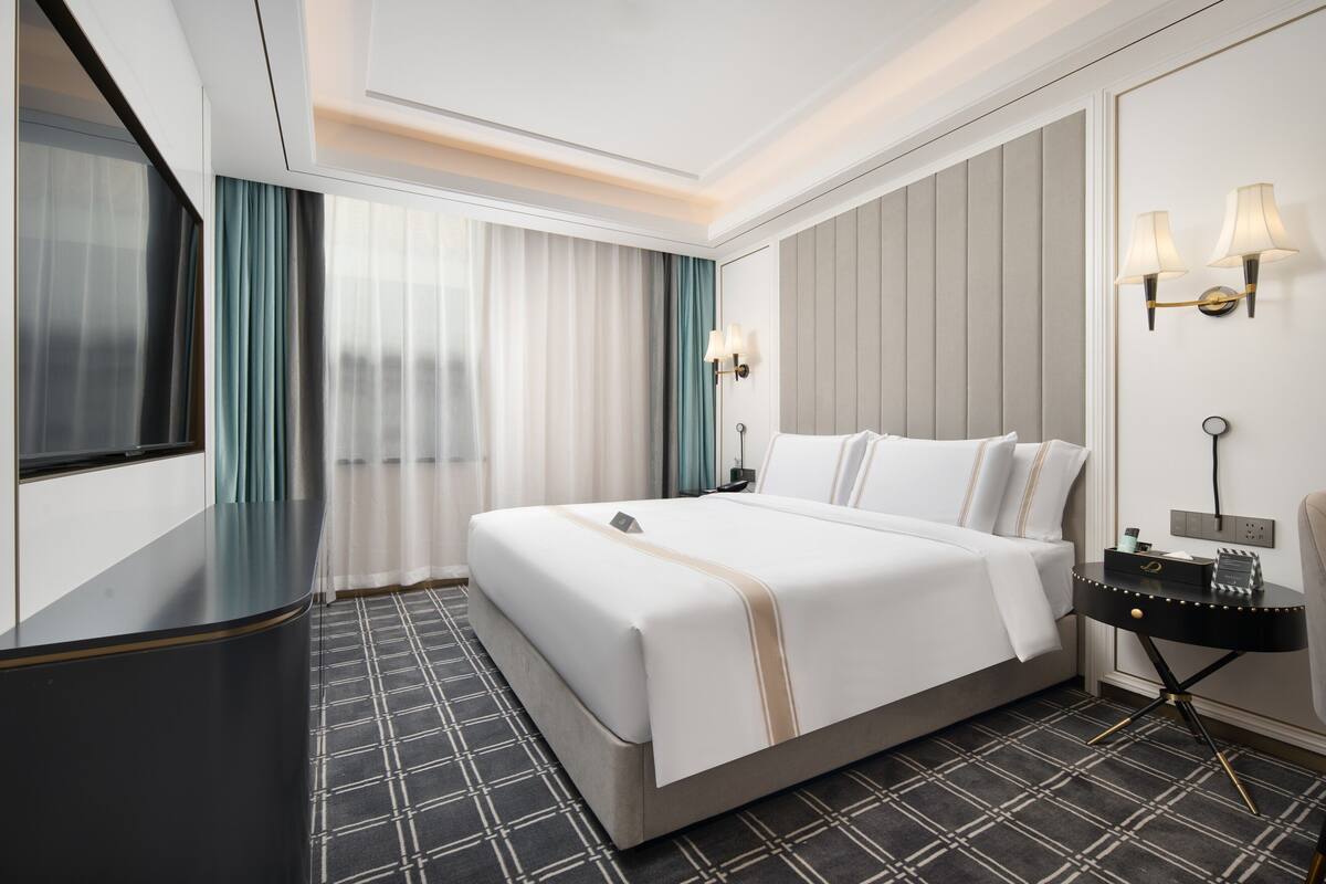 A hotel room with a bed, a nightstand, a lamp, a window with curtains, and a wall-mounted light fixture.