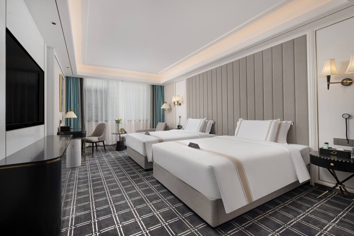 A hotel room with two beds, a desk, a chair, and a TV.