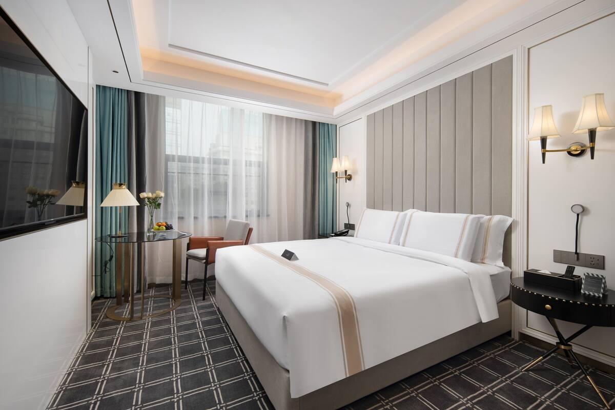 A hotel room with a large bed, a desk with a chair, a small table with a lamp, and a wall-mounted lamp.