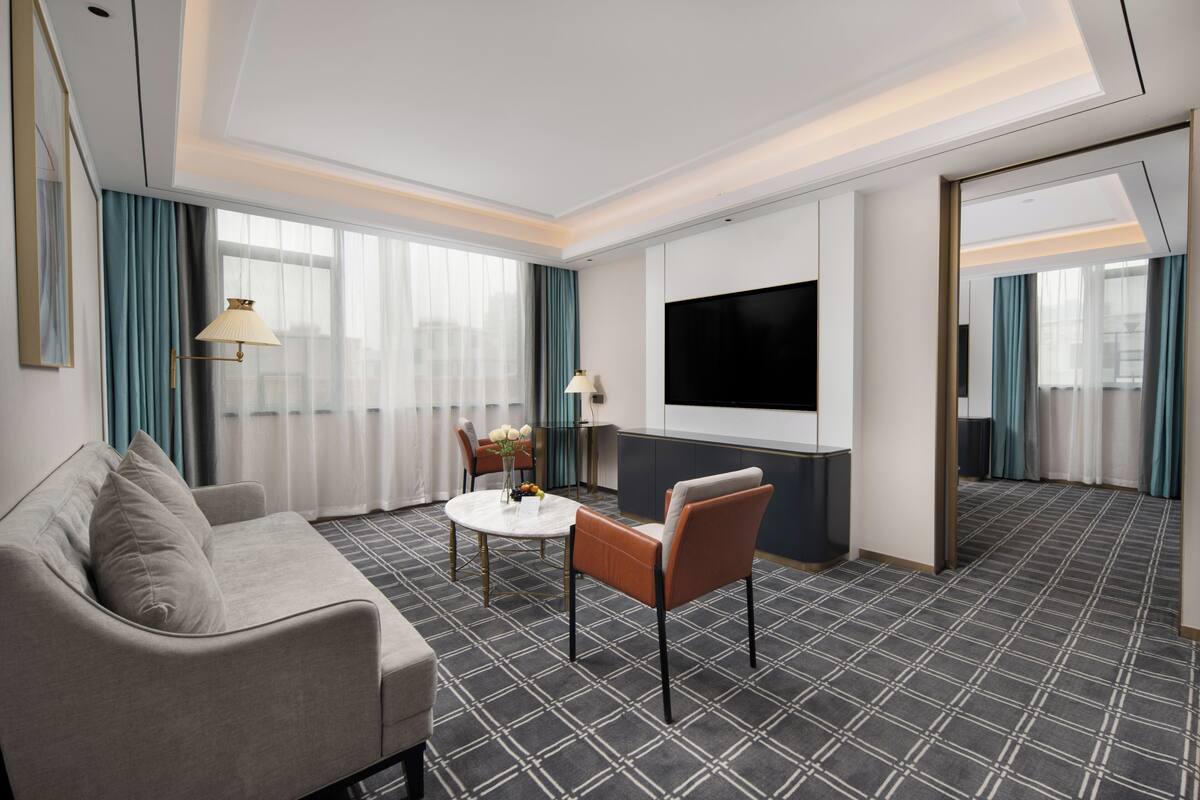 A hotel room with a sofa, a dining table, chairs, a TV, and a patterned carpet.