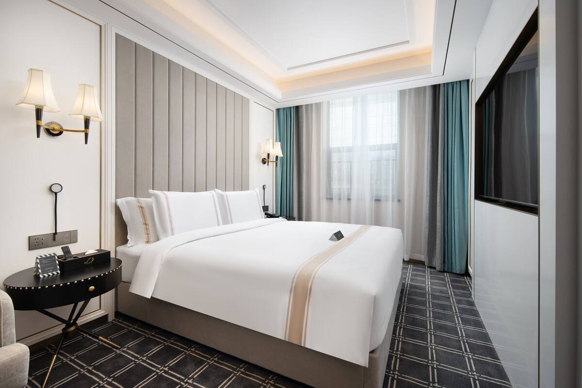 A hotel room with a large bed, two bedside lamps, a nightstand, and a view of the city through the window.
