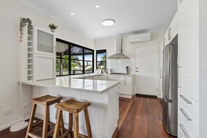 A modern kitchen with white cabinetry, a central island with stools, a stainless steel refrigerator, and a window with a view of greenery. - Relaxing Stay Near Brisbane’s Cultural Heart (Norman Park)