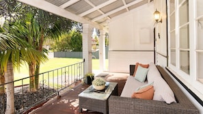 Eine überdachte Veranda mit einer Rattan-Couch, einem kleinen Tisch mit einer Pflanze und Blick auf eine grüne Rasenfläche und Palmen.