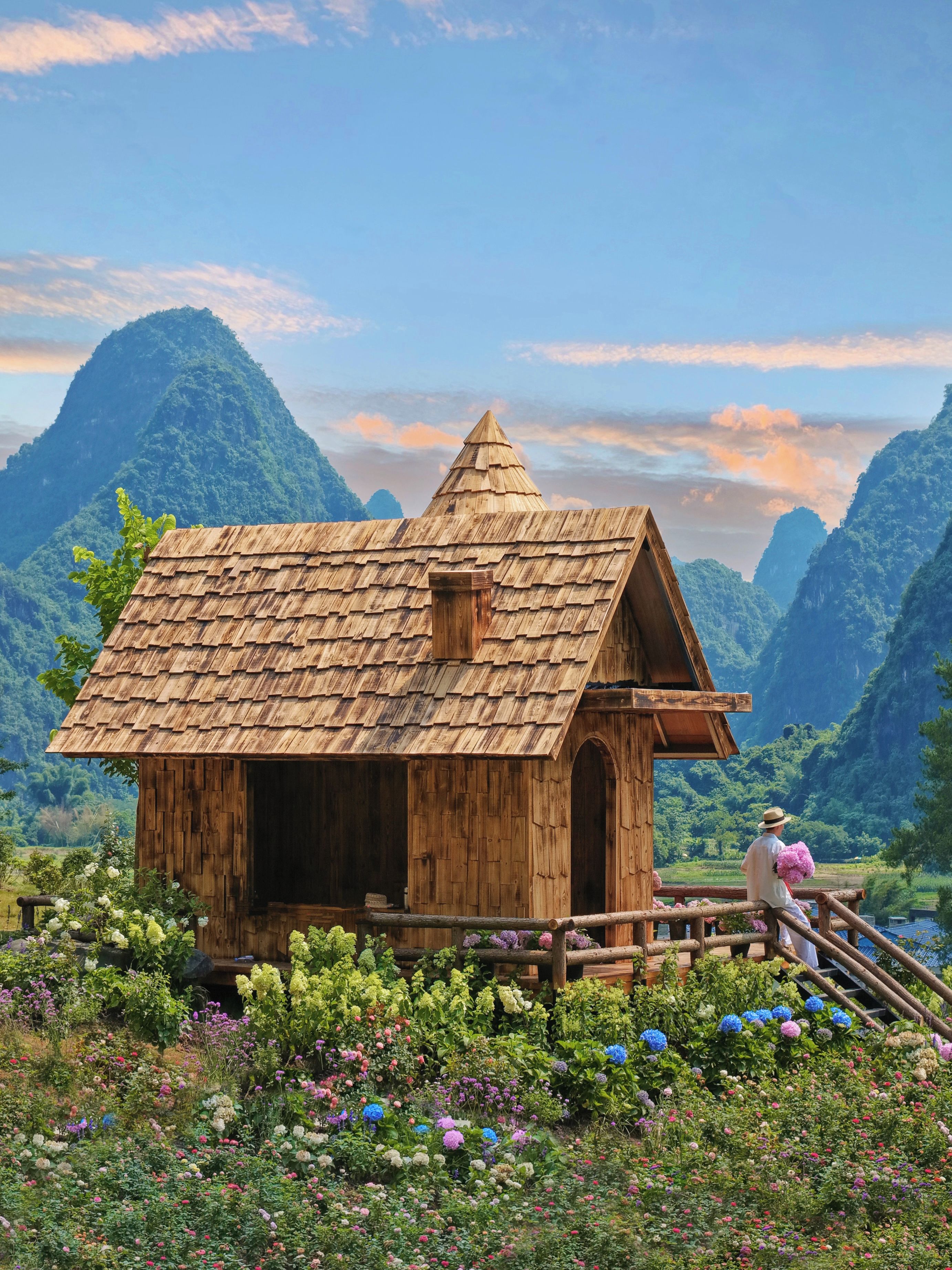 A wooden cabin with a sloped roof, surrounded by a variety of colorful flowers and set against a mountainous backdrop.