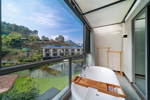 A modern bathroom with a large bathtub, a wooden tray, and a view of a residential area through a glass balcony. - FADAI Mountain Resort Yangshuo - The Art of Doing Nothing (Guilin)