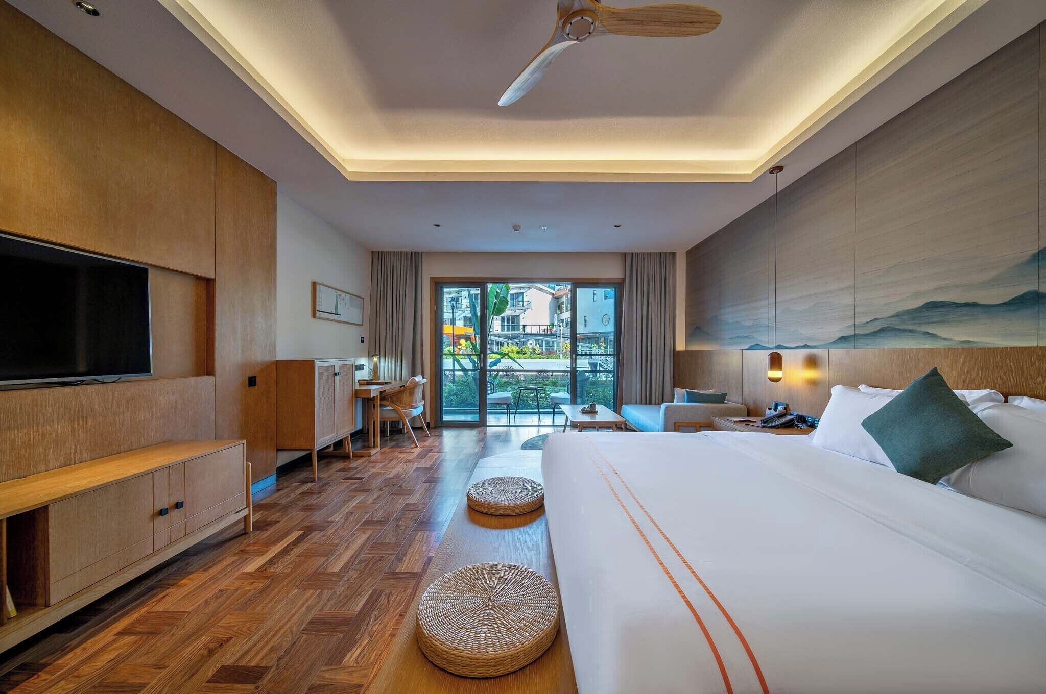 Habitación de hotel con cama, televisión, muebles de madera, ventilador de techo y vistas al exterior.