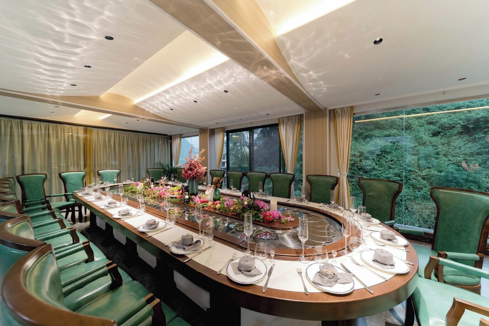 Une salle de conférence avec une table ronde, des chaises vertes et une vue sur une forêt, visible à travers de grandes fenêtres.