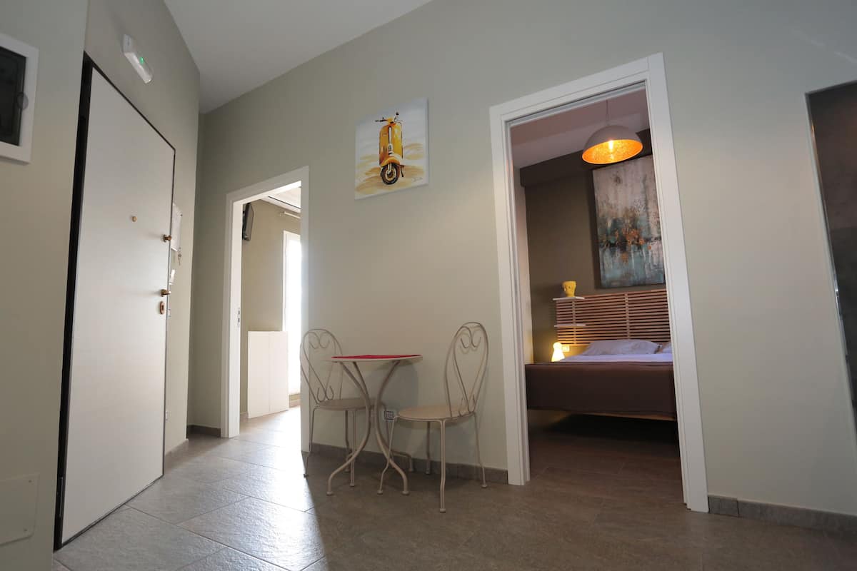 A hallway with a door, a small table with two chairs, a painting of a scooter, and a bed with a headboard and lamp.