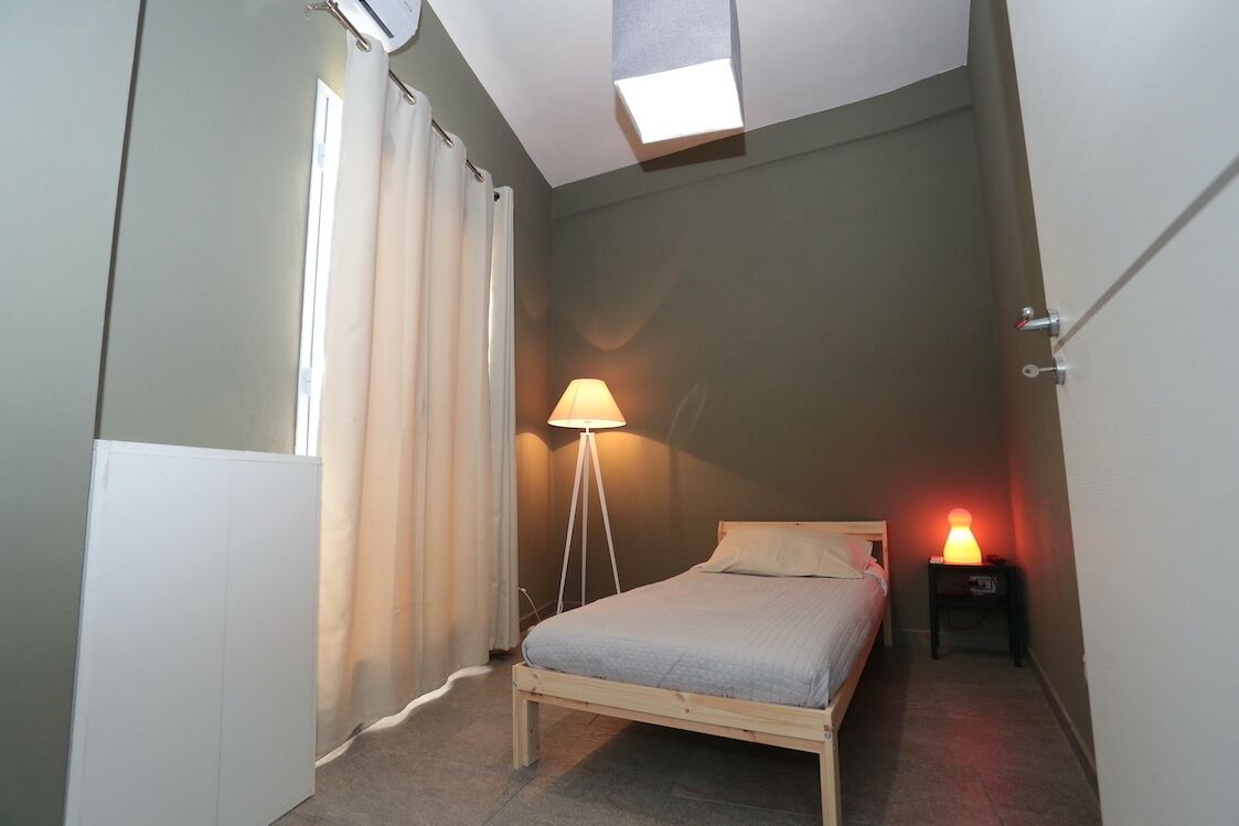 Ein Schlafzimmer mit einem Bett, einer Stehlampe, einem Nachttisch mit Lampe, einem Fenster mit Vorhängen und einer Deckenleuchte.