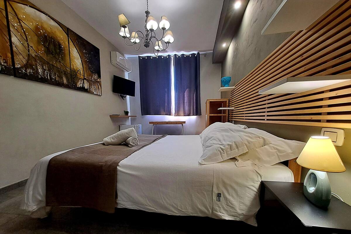 A modern hotel room with a bed, a bedside lamp, a TV, a painting on the wall, and a window with curtains.