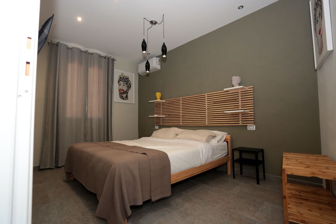 Une chambre à coucher comprenant un lit, une petite table, une étagère avec des objets décoratifs, un téléviseur fixé au mur et une fenêtre avec des rideaux.