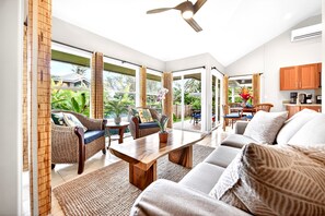 A spacious living area with a sofa, coffee table, and dining table, large windows with curtains, and a ceiling fan. - Honeymoon Cottage 1 Bed, 1 Bath with A/C, Steps to the Beach TVNC#5069 (Anahola)