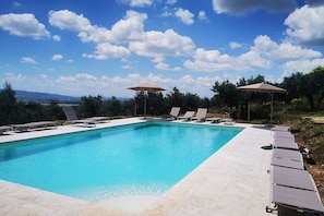 A rectangular swimming pool with clear blue water, surrounded by lounge chairs and umbrellas, set against a backdrop of trees and distant mountains under a partly cloudy sky.