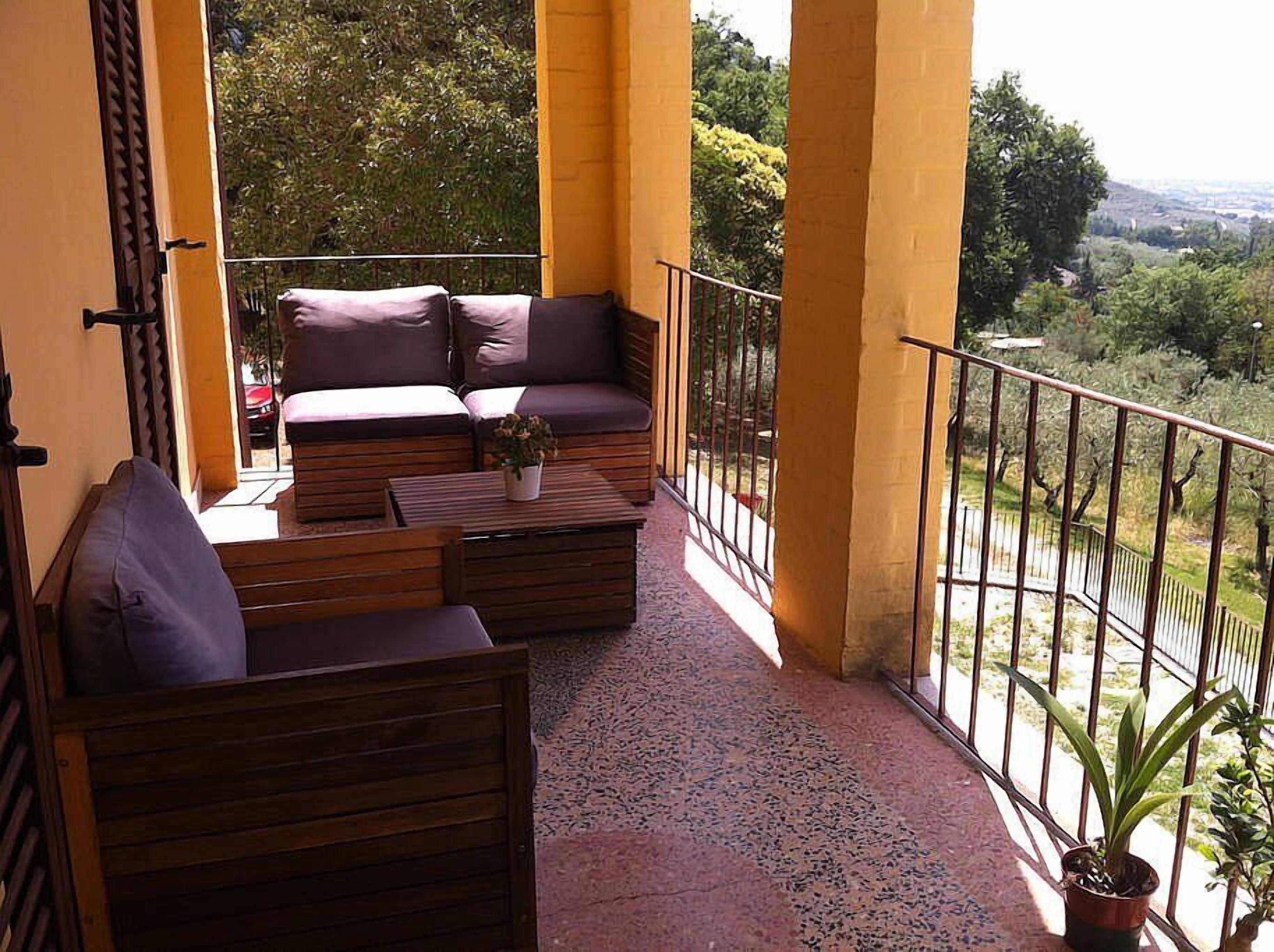 A balcony with a sofa and chairs, a small table with a plant, and a view of greenery and hills.