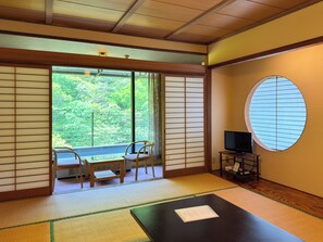 Ett traditionellt japanskt rum med tatamimattor på golvet, en skjutdörr som leder ut till en balkong, en tv på ett stativ och ett runt fönster.