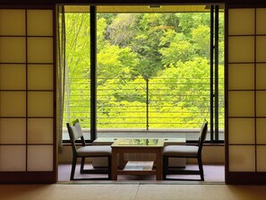 A room with tatami flooring, a low wooden table, two chairs, and a large window with a view of a green forest. - HIRANOYA (Takayama)