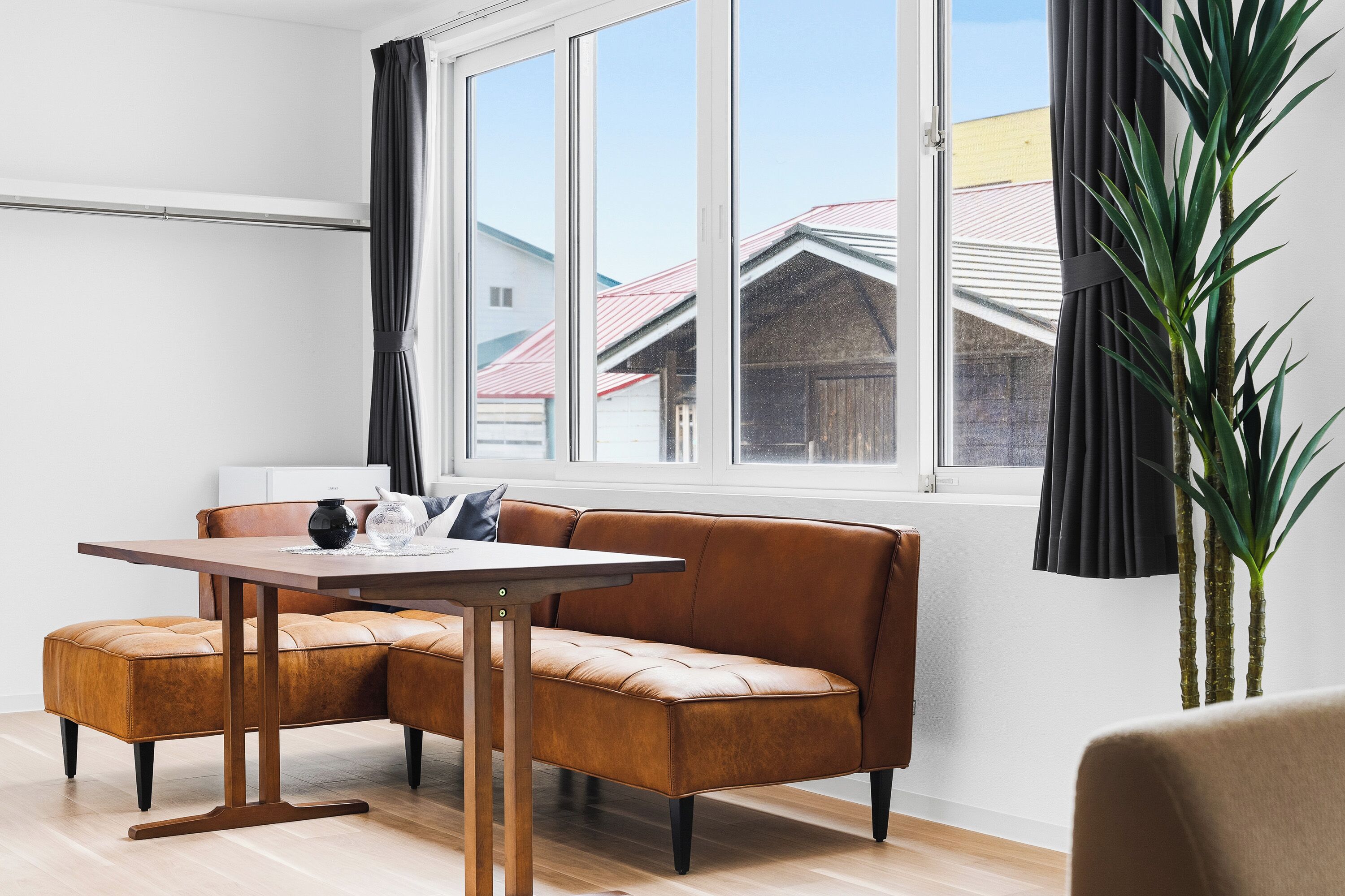 A modern room with a wooden dining table, leather sofa, and a large window with curtains.