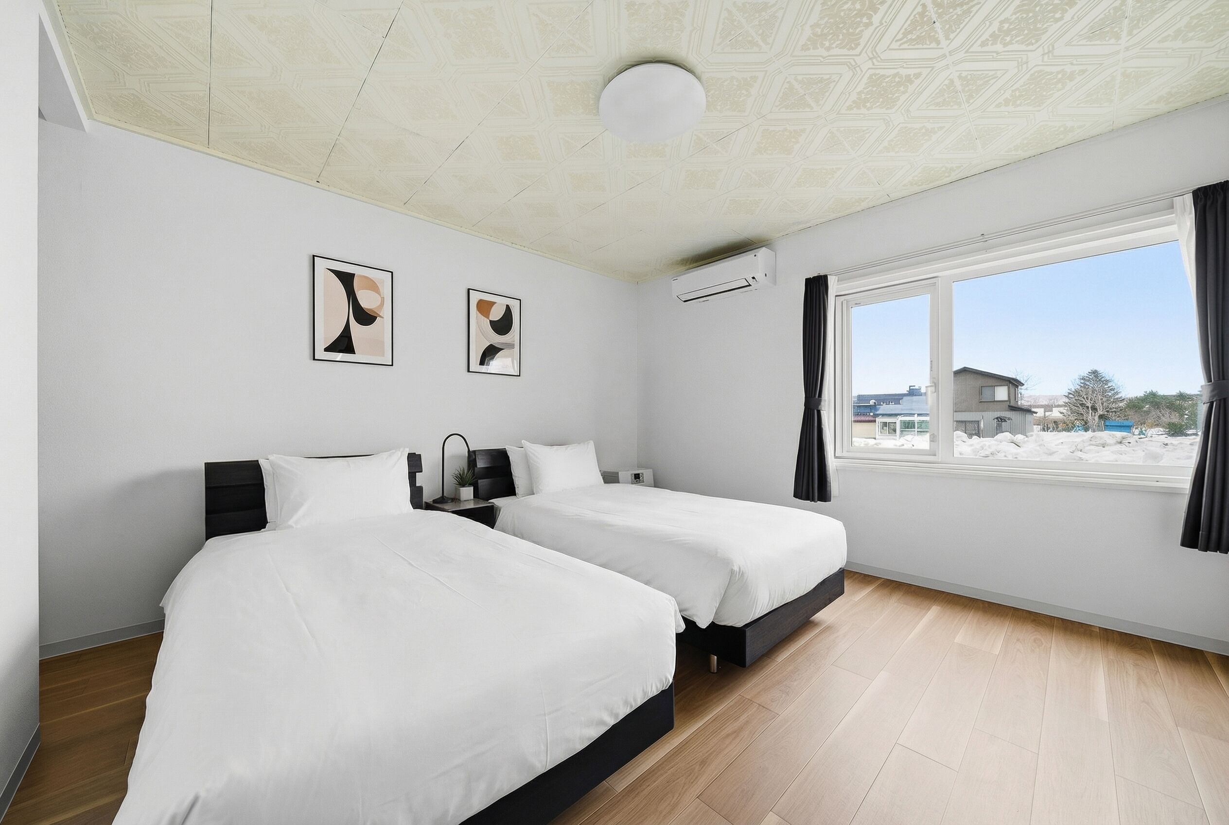 A hotel room with two beds, a ceiling with a geometric pattern, an air conditioning unit, and a window with a view of buildings.