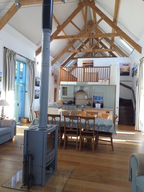 A spacious living area with a high ceiling, wooden beams, a central fireplace, a dining table with chairs, and a kitchen area with a staircase.
