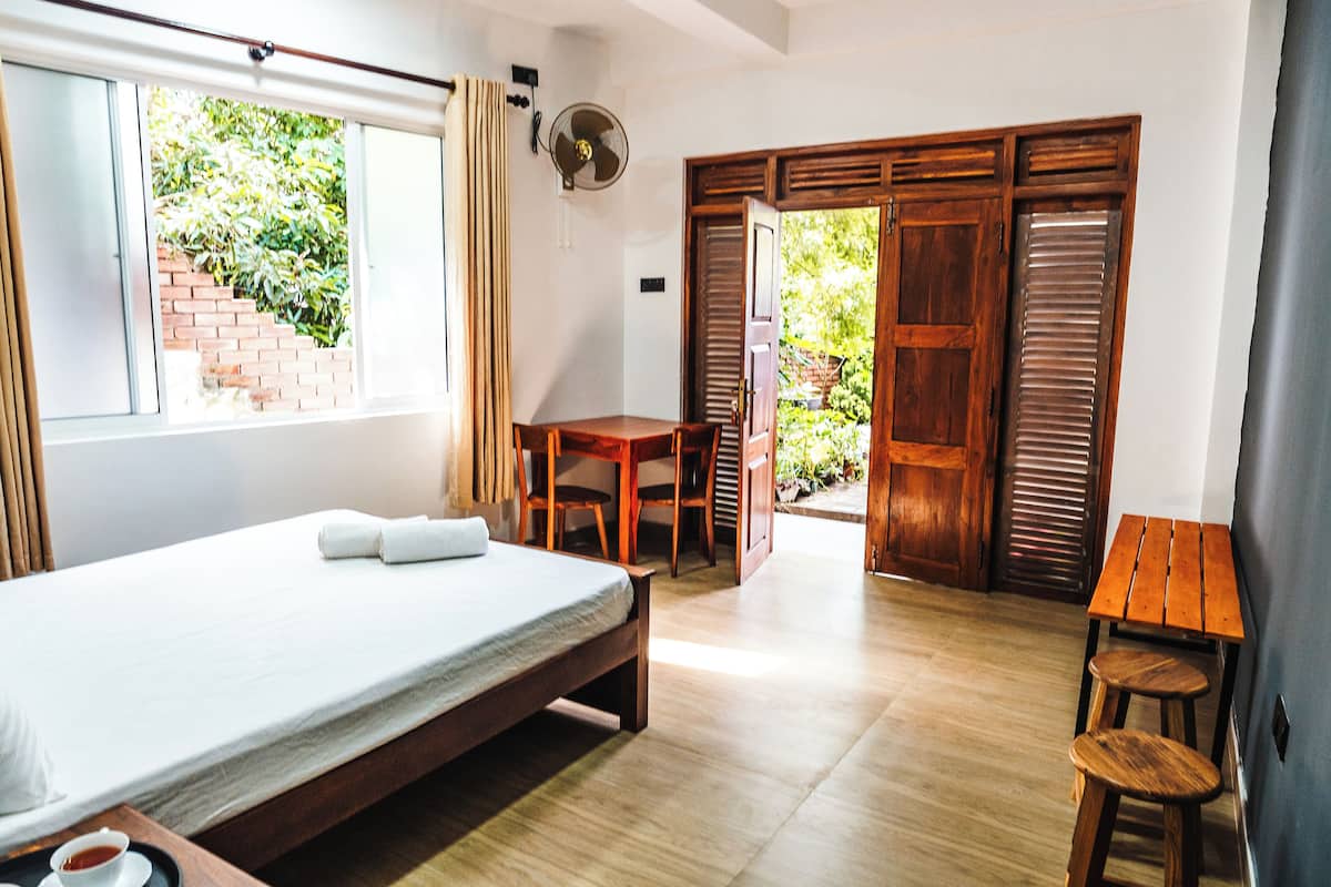 A bedroom with a bed, a table, and a chair. There is a window and a door leading to an outdoor area.