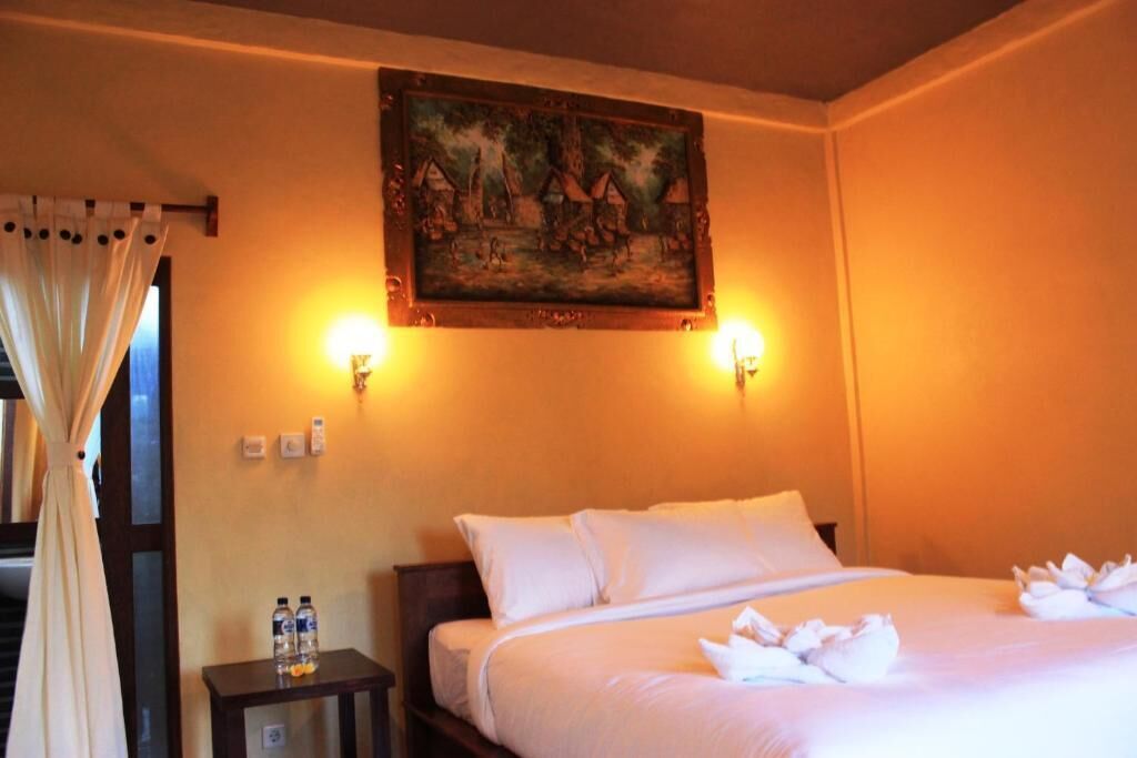 Habitación de hotel con cama, un cuadro en la pared, dos apliques, una mesita de noche con botellas y una cortina.