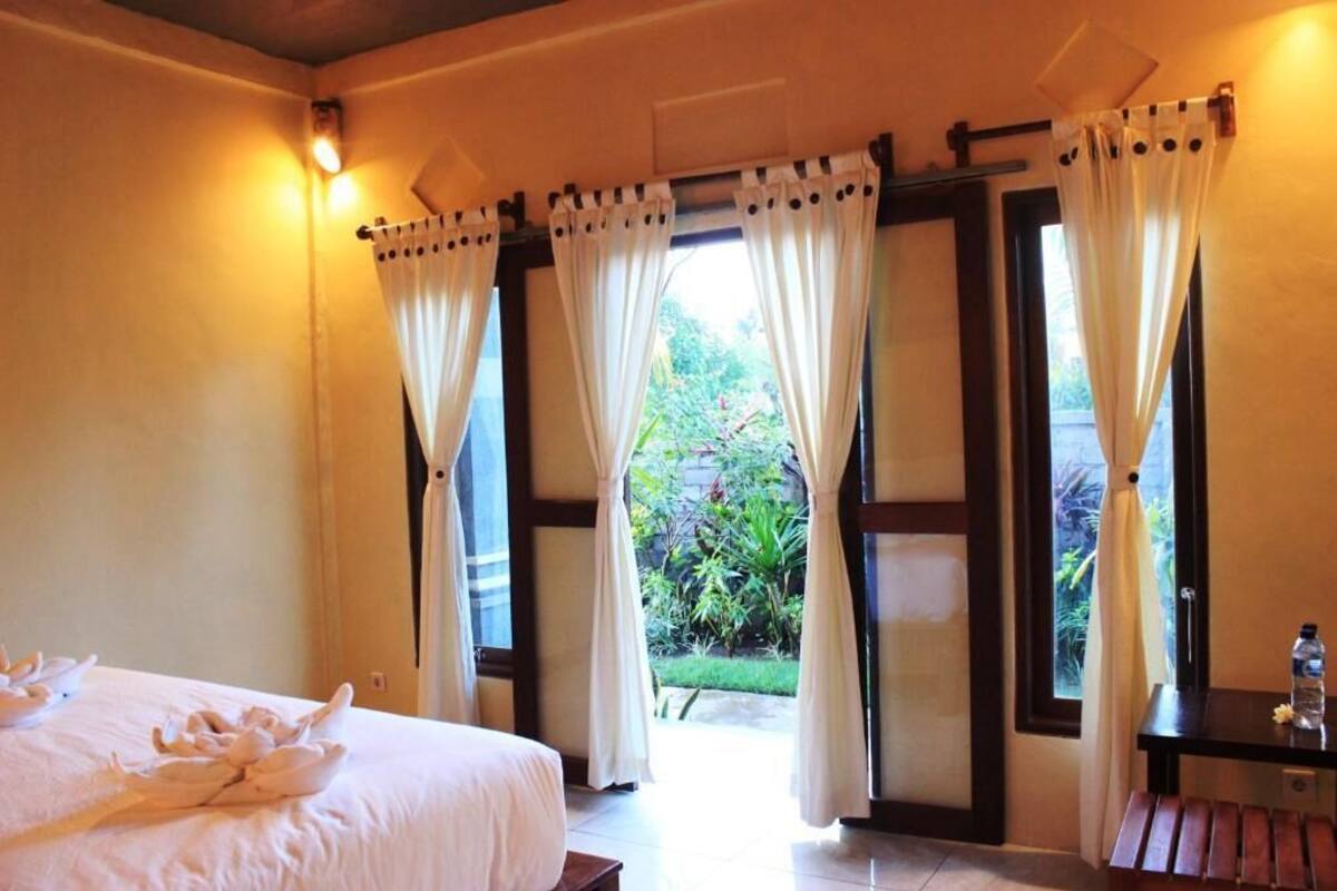 A hotel room with a bed, curtains, a view of greenery, and a small table with a bottle.