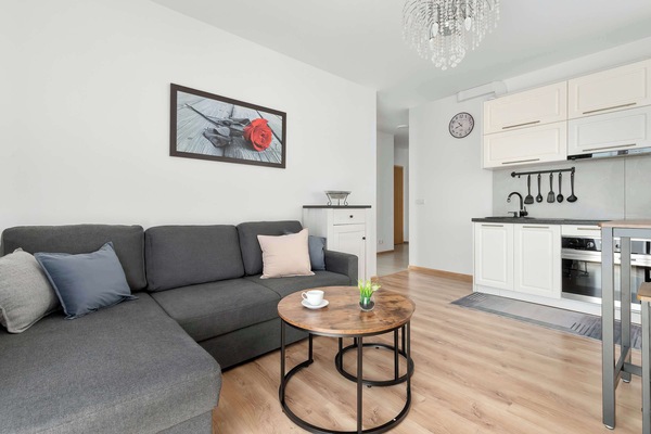 A spacious living room features a large, dark L-shaped couch paired with two round, industrial-style coffee tables. The space connects to a modern kitchen with white cabinets, while daylight from a large window makes the interior bright and inviting.