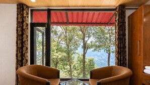 Um quarto com duas poltronas marrons, uma mesa de vidro e vista para árvores através de uma grande janela.