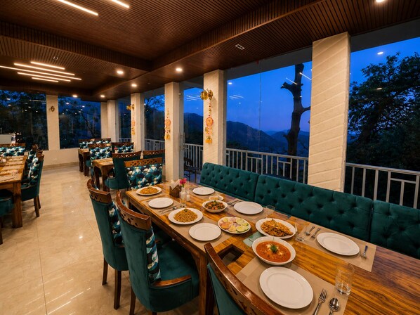 A restaurant with a long wooden table, teal chairs, and a view of the mountains. - Kavin Highland - The Mall (Dalhousie)