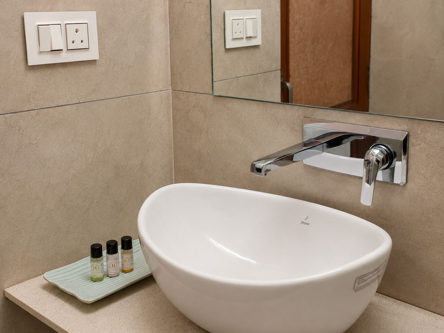 Un bagno con un lavabo a incasso bianco, un rubinetto cromato e uno specchio sopra. Accanto al lavabo, ci sono tre piccole bottiglie su un vassoio.