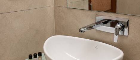 Un bagno con un lavabo a incasso bianco, un rubinetto cromato e uno specchio sopra. Accanto al lavabo, ci sono tre piccole bottiglie su un vassoio.