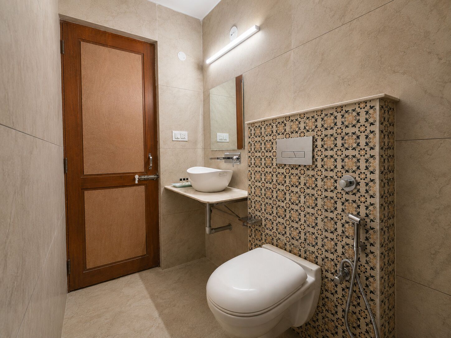 Baño con puerta de madera, lavamanos blanco, inodoro y ducha con revestimiento estampado.