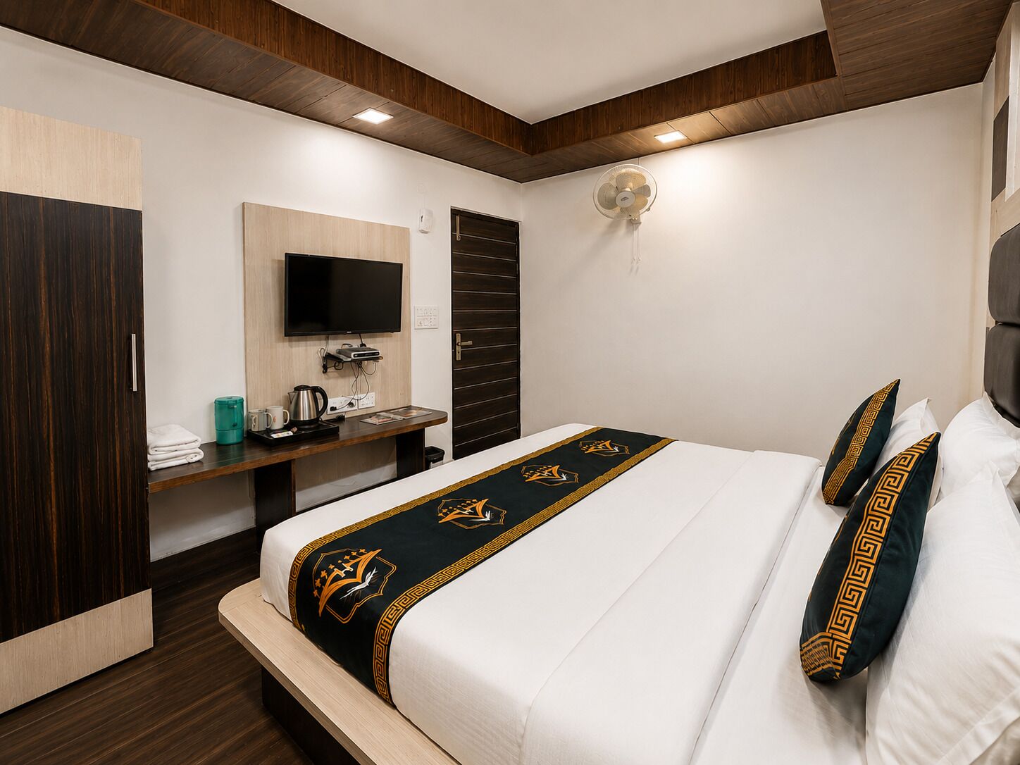 Una camera d'hotel con un letto, una TV, una scrivania con un bollitore e una ventola a muro.