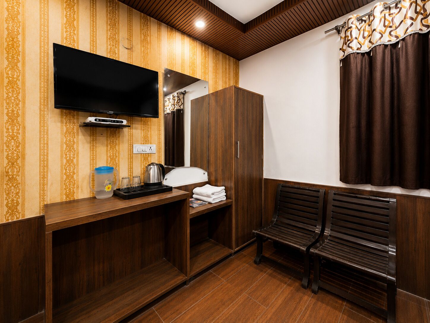Ein Hotelzimmer mit einem Holzschrank, einem Flachbildfernseher, einem kleinen Tisch mit Wasserkocher, Gläsern und einem Wasserspeicher. Zwei Holzbänke und ein mit Vorhängen versehenes Fenster sind ebenfalls vorhanden.