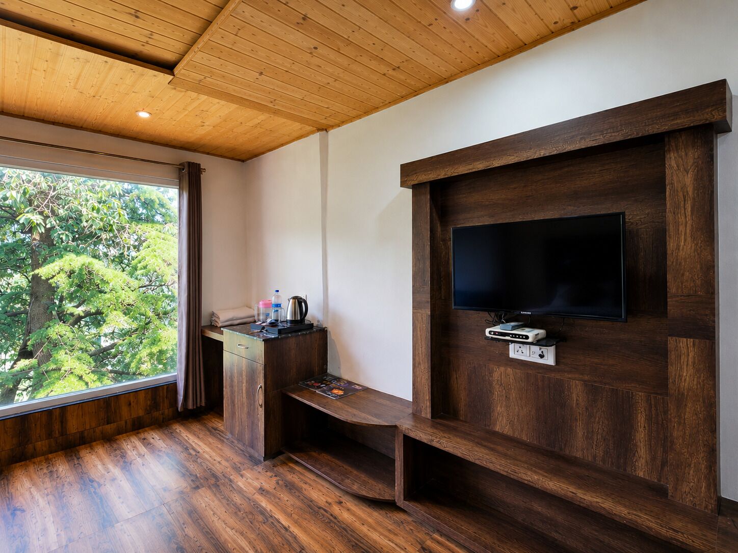 A room with a wooden ceiling, a TV mounted on a wooden wall, a desk with a kettle, and a window with a view of trees.
