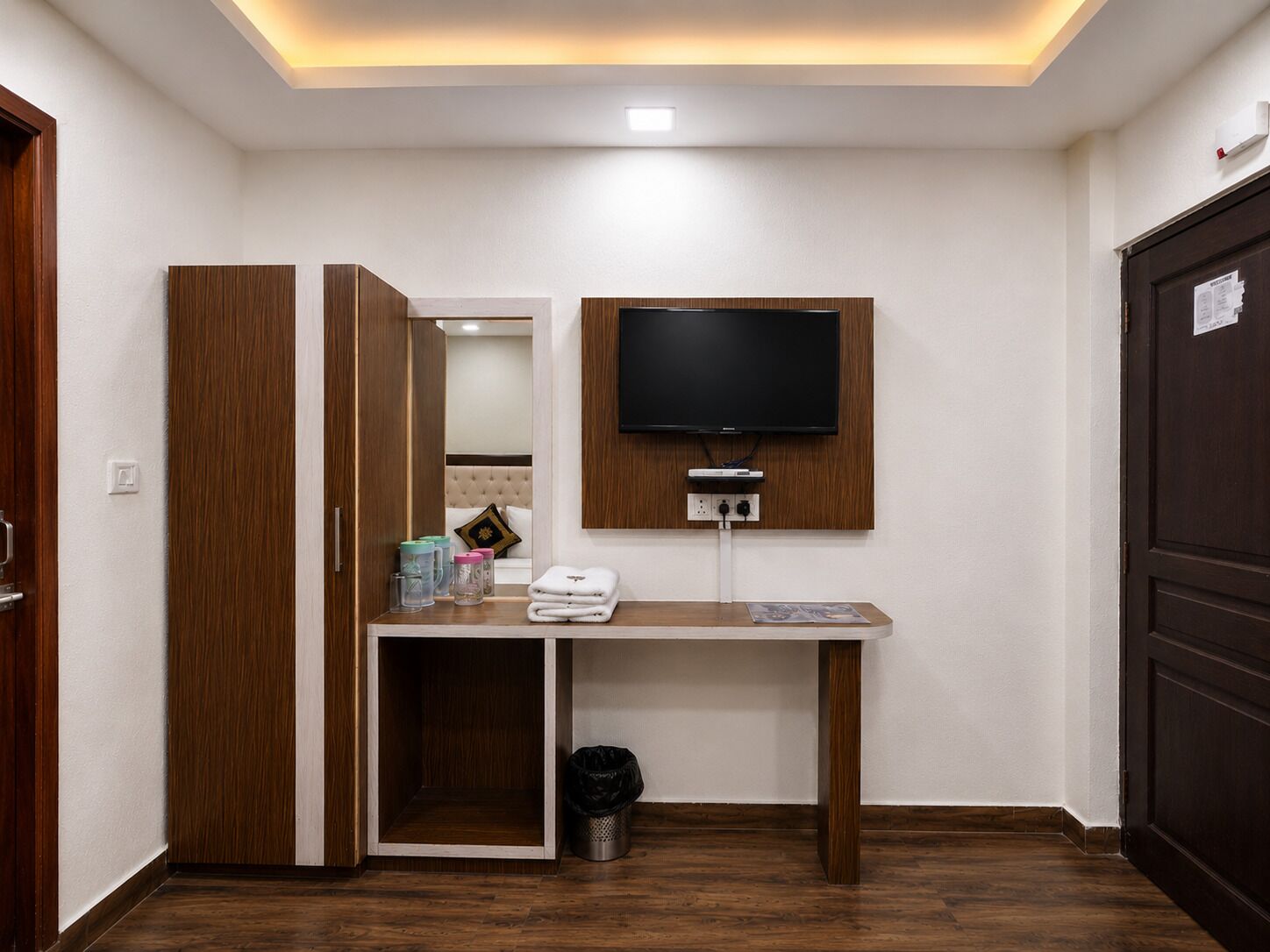 Habitación de hotel con armario de madera, televisor de pantalla plana en la pared, escritorio pequeño con cesto de basura y cama con cabecero.