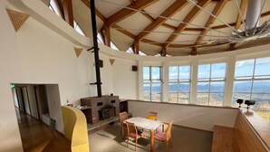A spacious room with a high ceiling, a fireplace, a dining area, and large windows offering a view of the landscape. - Lodge Cowbell (Takko)