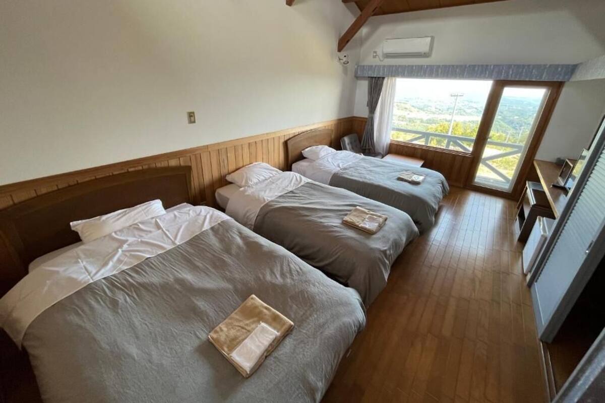 A room with four beds, wooden floors, a window with a view, and a ceiling fan.